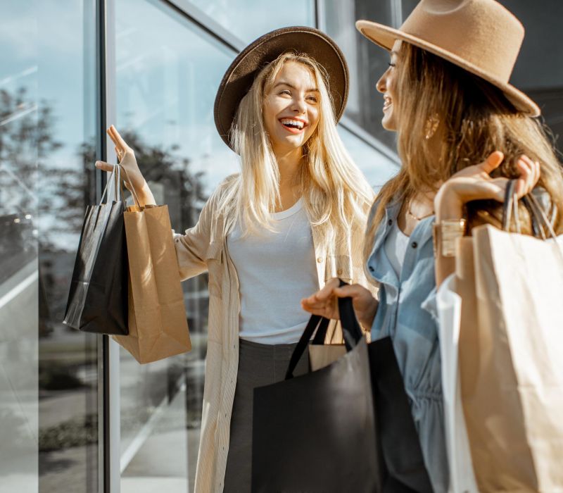 Two women smiling and holding shopping bags while walking outdoors by glass windows.