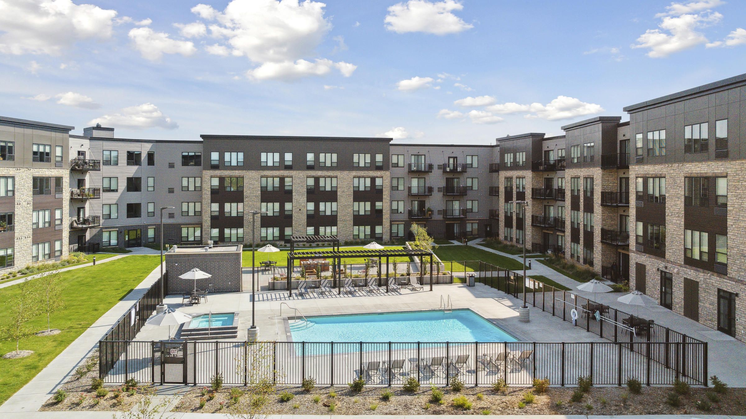 Modern apartment complex with a central fenced pool area, lounge chairs, and clear sky with clouds above.