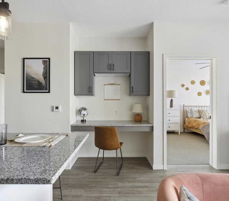 Modern apartment interior with a small desk workspace, gray cabinets, and a view into a cozy bedroom.