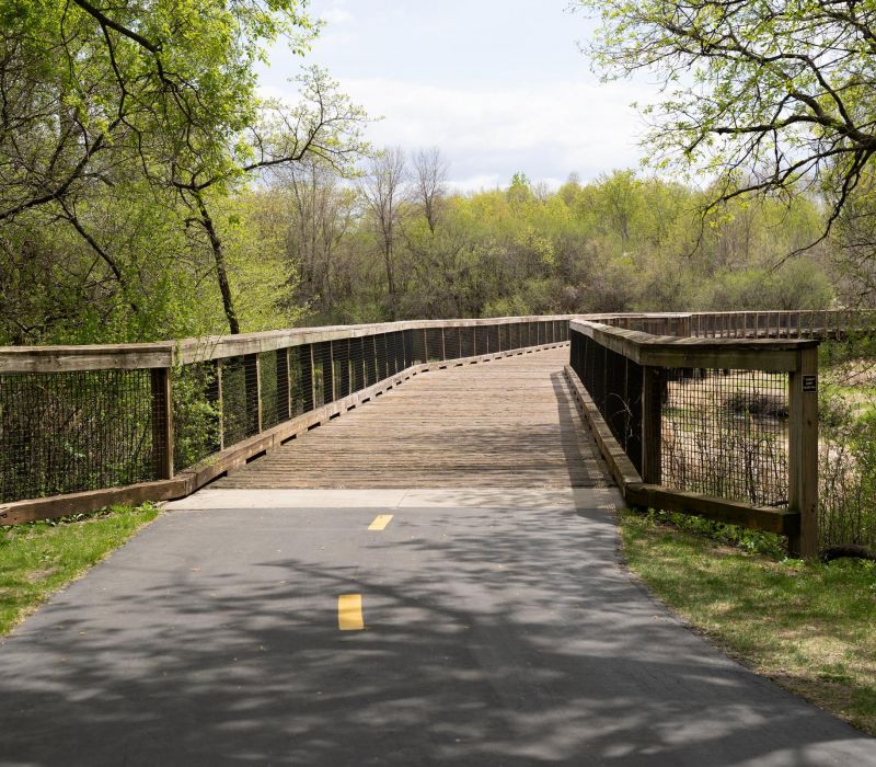 A paved trail leads to a wooden footbridge surrounded by green trees and foliage on a sunny day.