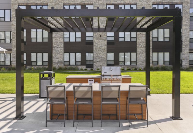 Outdoor patio with a pergola, bar seating, grill, and modern building in the background.