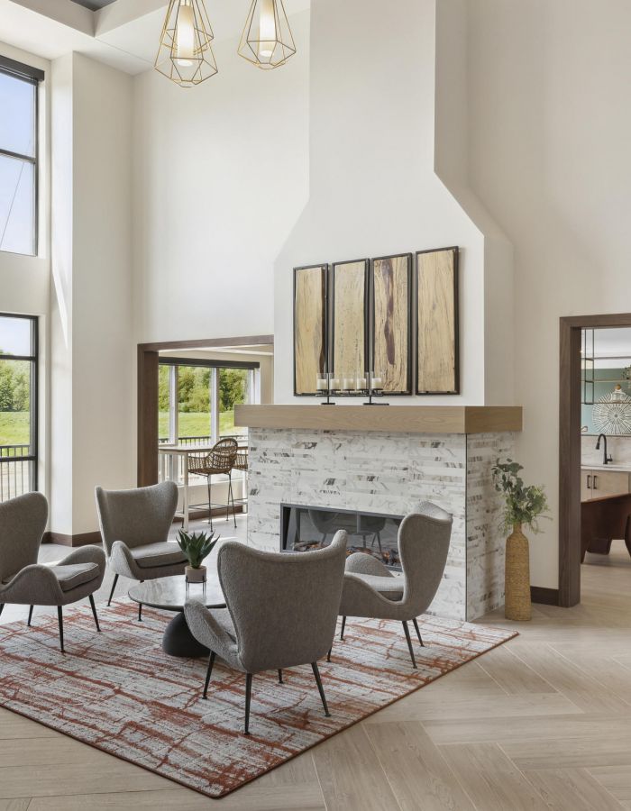 Modern living room with large windows, gray chairs around a rug, and a fireplace; view into another room.