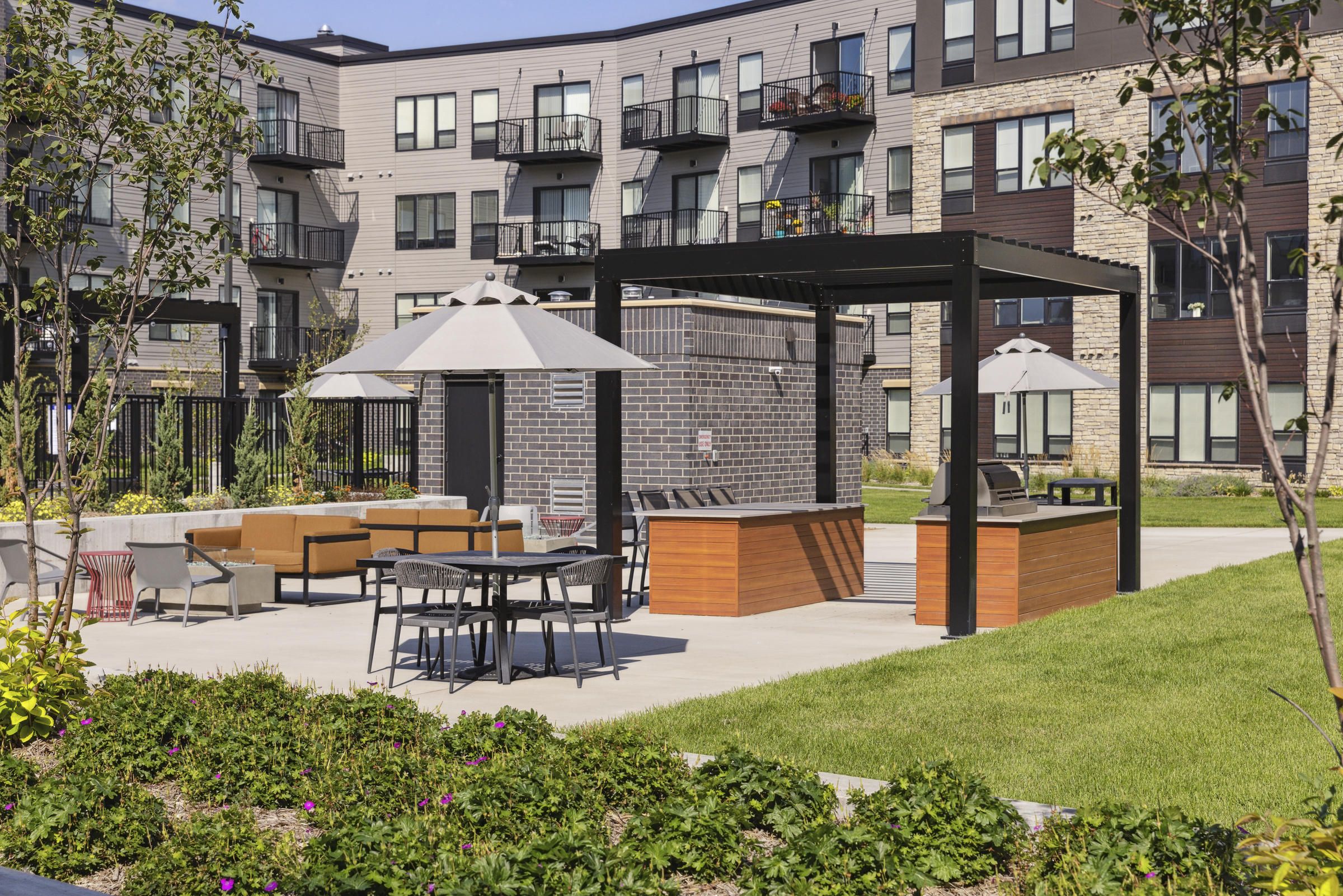 Outdoor lounge area with tables, umbrellas, grills, and seating, in front of a modern apartment building.