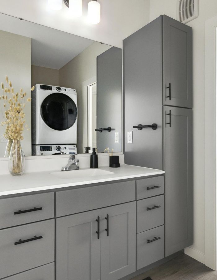 Modern bathroom with gray cabinets, a large mirror, and a stacked washer-dryer reflected in the mirror.