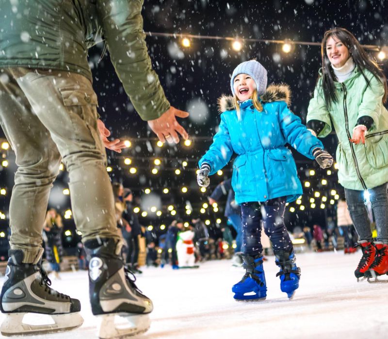 A child ice skates outdoors with two adults, smiling under string lights and gently falling snow.