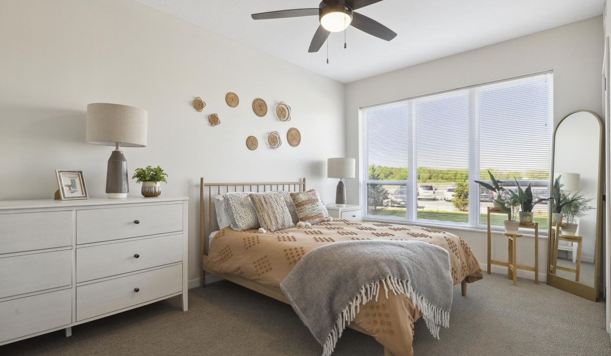 Bright bedroom with a bed, white dresser, wall art, plants, and large window letting in natural light.