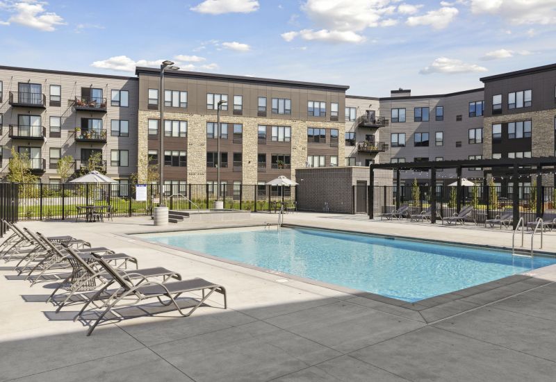 Modern apartment complex with balconies, outdoor pool, lounge chairs, and pergola on a sunny day.