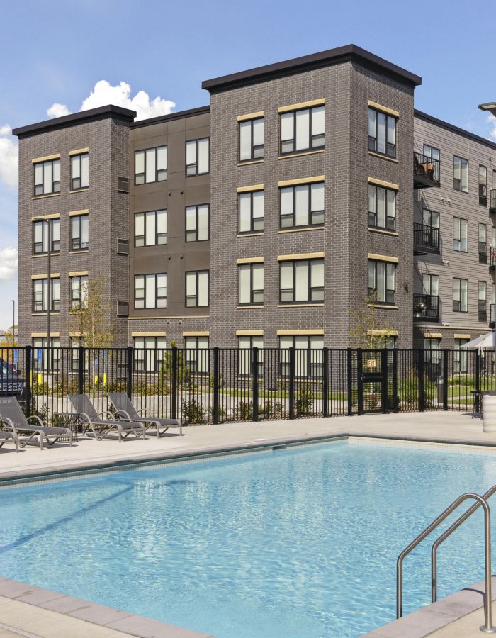 A modern apartment building with a gated outdoor swimming pool and lounge chairs under a clear sky.