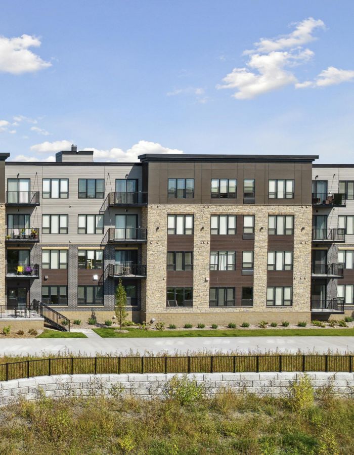 Modern four-story apartment building with balconies, large windows, and landscaped entrance under a blue sky.