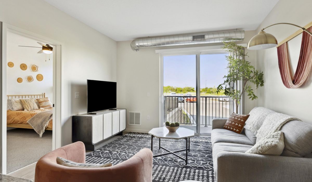 Modern living room with a sofa, TV, and large window; bedroom visible through an open door.